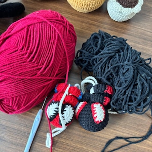 May include: A large ball of red yarn and a large ball of black yarn positioned next to a light blue crochet hook. Two small crocheted figures are present, one featuring red, blue, and white details, and the other featuring black, red, and white.