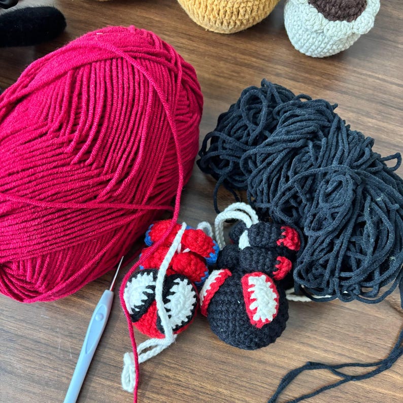 May include: A large ball of red yarn and a large ball of black yarn positioned next to a light blue crochet hook. Two small crocheted figures are present, one featuring red, blue, and white details, and the other featuring black, red, and white.