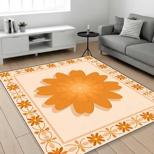 May include: A rectangular area rug with a large orange flower design in the center. The rug has a light peach background with an orange floral border. The rug is in a living room setting with a gray sofa and white cabinet.