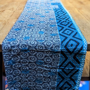 May include: A blue and white patterned table runner with a floral design on one side and a geometric pattern on the other. The runner is made of fabric and is displayed on a wooden table. The runner is approximately 183 cm long.