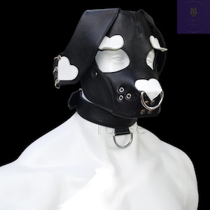 Könnte beinhalten: Eine schwarze Welpenmaske aus lederähnlichem Material mit weißen herzförmigen Akzenten an Ohren und Nase. Die Maske verfügt über Augenausschnitte, silberfarbene Metallösen, einen Nasenring sowie ein passendes Halsband mit silbernem D-Ring.