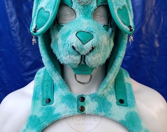 May include: A turquoise and white tie-dye faux fur hood with long rabbit ears, featuring heart-shaped accents, a silver chain across the forehead, and a metal D-ring at the chin. The hood is attached to a matching chest harness with black snaps.