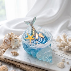 Puede incluir: Un portavelas azul en forma de concha con una cola de sirena, estrellas de mar y conchas. La vela está encendida y el portavelas se encuentra sobre una bandeja de mármol con conchas y coral adicionales. La etiqueta dice "Ocean Soul Handmade".