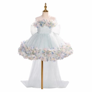 Puede incluir: Un vestido formal azul claro con una falda de tul y una larga cola. El corpiño está adornado con detalles florales en colores pastel, y hay un gran lazo en la espalda. El vestido se exhibe en un maniquí dorado.