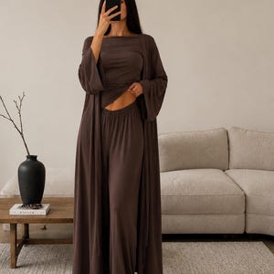 Op de afbeelding: Een donkerbruine driedelige loungeset bestaande uit een lang vest met open voorzijde en lange mouwen, een top met korte mouwen en ronde hals, en een broek met hoge taille en wijde pijpen van gladde stof.