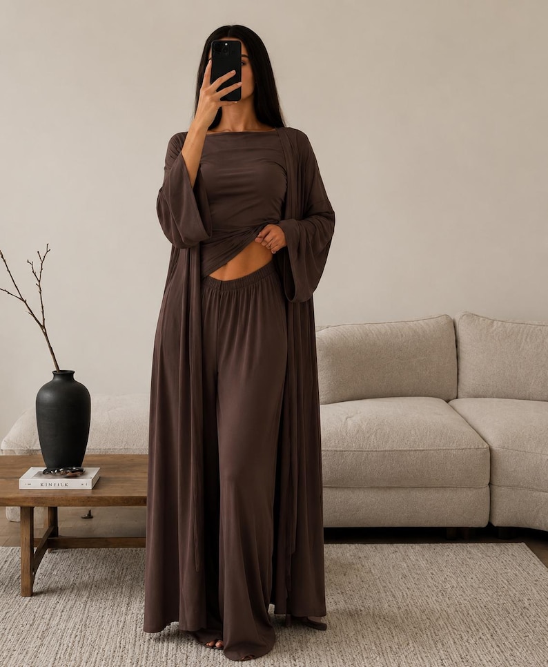 K&ouml;nnte beinhalten: Ein dunkelbraunes dreiteiliges Lounge-Set, bestehend aus einer lang&auml;rmeligen, offenen Strickjacke, einem kurz&auml;rmeligen Oberteil mit Rundhalsausschnitt und einer hoch taillierten Hose mit weitem Bein aus glattem Stoff.
