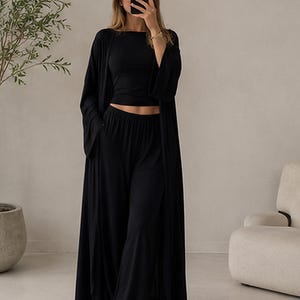 K&ouml;nnte beinhalten: Ein schwarzes dreiteiliges Lounge-Set bestehend aus einem bauchfreien Kurzarm-Oberteil, einer hoch taillierten Hose mit weitem Bein und elastischem Bund sowie einer langen, vorne offenen Strickjacke mit weiten &Auml;rmeln aus schwarzem Stoff.