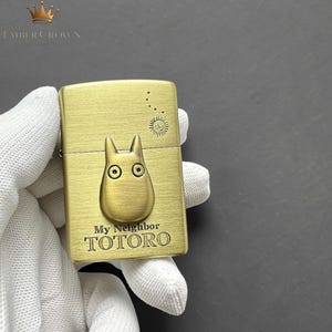 Puede incluir: Un encendedor dorado con una imagen en relieve de Totoro de "Mi Vecino Totoro". El encendedor tiene el texto "My Neighbor TOTORO" grabado en la parte delantera. La esquina superior derecha tiene un dise&ntilde;o de sol y puntos.