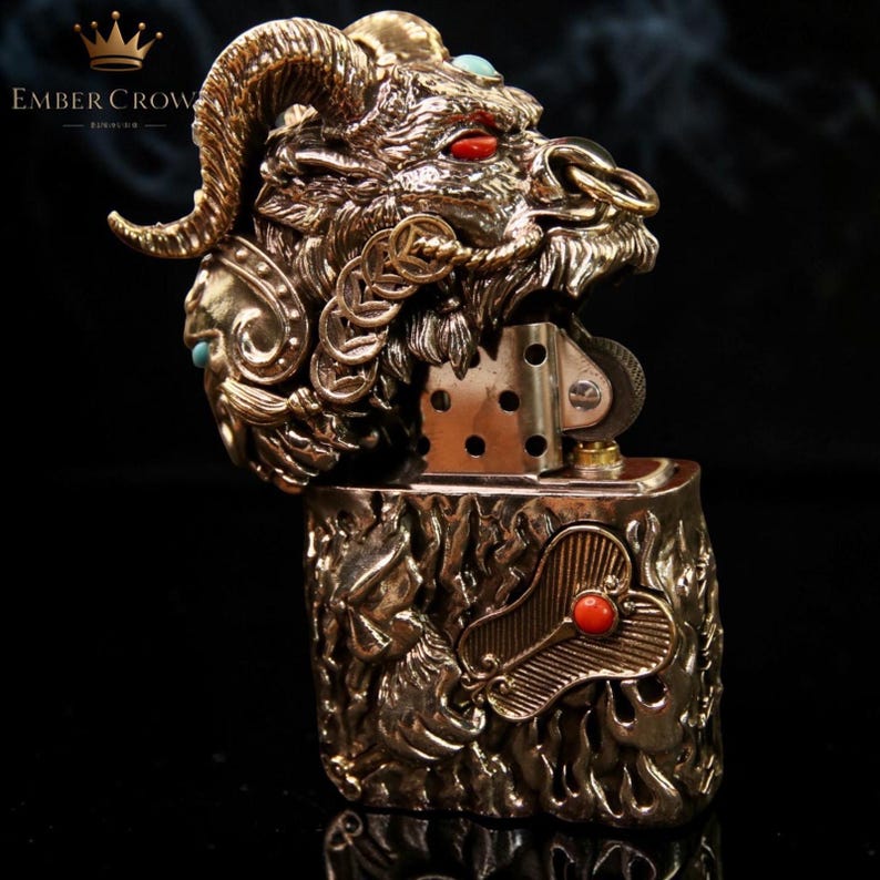 K&ouml;nnte beinhalten: Ein bronzefarbenes Metallfeuerzeug mit einem Widderkopf-Design. Der Widderkopf hat rote Augen und t&uuml;rkisfarbene Akzente. Das Feuerzeug hat aufwendige Details und ein dekoratives Design am K&ouml;rper. Der Markenname "Ember Crow" ist sichtbar.