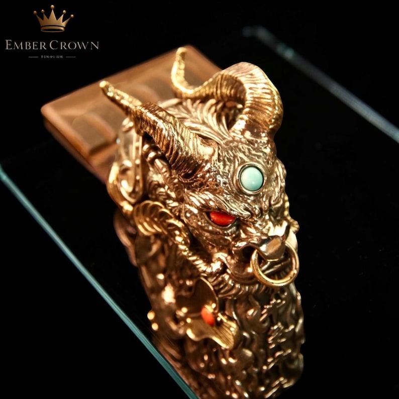 K&ouml;nnte beinhalten: Eine goldfarbene, verzierte Drachenkopfskulptur mit roten Augen und einem t&uuml;rkisfarbenen Stein. Der Drache hat gebogene H&ouml;rner und einen Ring im Maul. Die Skulptur steht auf einem rechteckigen Sockel. Der Text "EMBER CROWN" befindet sich oben links.