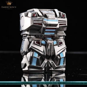 May include: A silver-toned, geometric art deco style lighter with red and blue enamel accents. The lighter has a rectangular shape with a detailed, layered design. The Ember Crown logo is visible in the top left corner.