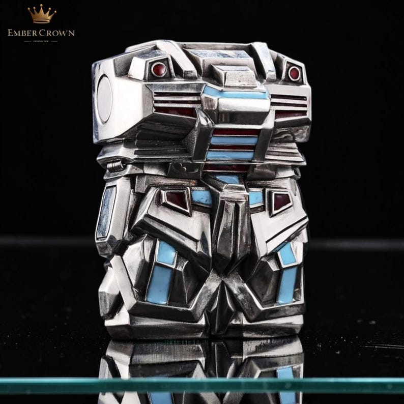 May include: A silver-toned, geometric art deco style lighter with red and blue enamel accents. The lighter has a rectangular shape with a detailed, layered design. The Ember Crown logo is visible in the top left corner.