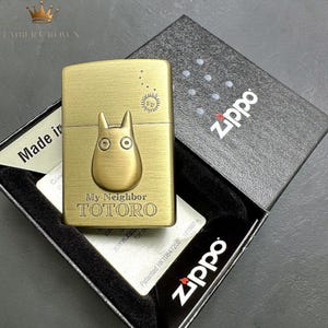 Puede incluir: Un encendedor Zippo de color lat&oacute;n con un dise&ntilde;o de Totoro de "Mi Vecino Totoro". El encendedor est&aacute; en su caja negra original con el logotipo de Zippo. El dise&ntilde;o incluye el texto "My Neighbor TOTORO" y un sol.