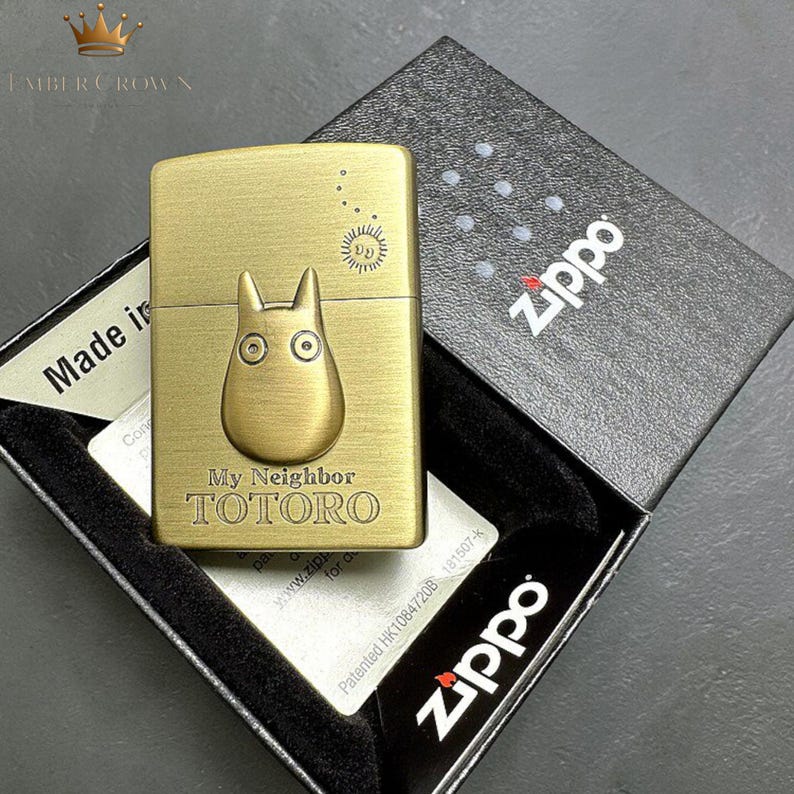 Puede incluir: Un encendedor Zippo de color lat&oacute;n con un dise&ntilde;o de Totoro de "Mi Vecino Totoro". El encendedor est&aacute; en su caja negra original con el logotipo de Zippo. El dise&ntilde;o incluye el texto "My Neighbor TOTORO" y un sol.