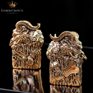 K&ouml;nnte beinhalten: Zwei verzierte, goldfarbene Metallskulpturen von Widderk&ouml;pfen mit gebogenen H&ouml;rnern, roten Augen und t&uuml;rkisen und roten Akzenten. Die Skulpturen weisen komplizierte Details auf und sind vor einem dunklen Hintergrund platziert. Der Text "EMBER CROWN" befindet sich oben links.