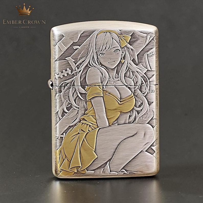 Puede incluir: Un encendedor de metal plateado con una ilustraci&oacute;n grabada de estilo anime de una mujer con un vestido amarillo. El encendedor tiene un acabado de metal cepillado y la marca "EMBER CROWN" es visible en la parte superior izquierda.