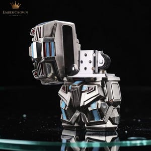 May include: A silver-colored lighter with a geometric design, featuring blue and red accents. The lighter is open, revealing the internal components. The brand name "EMBER CROWN" is visible in the top left corner.