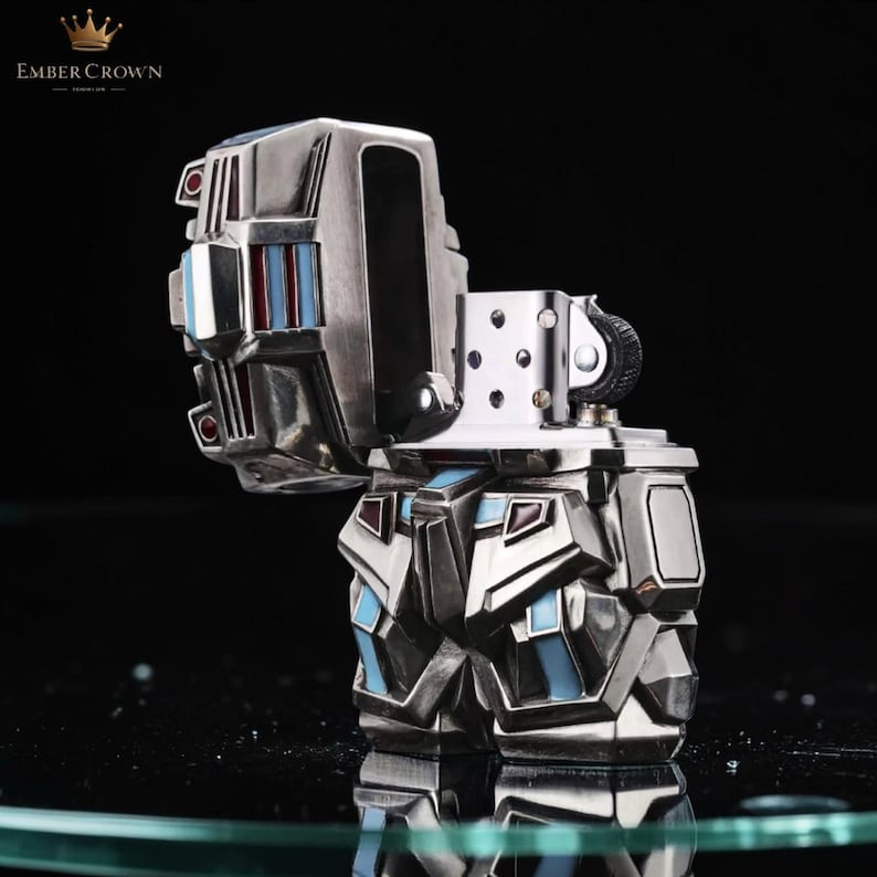 May include: A silver-colored lighter with a geometric design, featuring blue and red accents. The lighter is open, revealing the internal components. The brand name "EMBER CROWN" is visible in the top left corner.