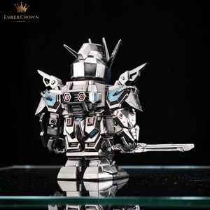 May include: A silver-colored robot figurine with a sword. The robot has a detailed design with blue and red accents. The Ember Crown logo is visible in the upper left corner.