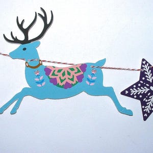May include: A decorative garland featuring a light blue reindeer cutout with black antlers and a colorful floral saddle, flanked by a red star cutout with white leaf patterns and a dark purple star cutout with white snowflake designs on a string.