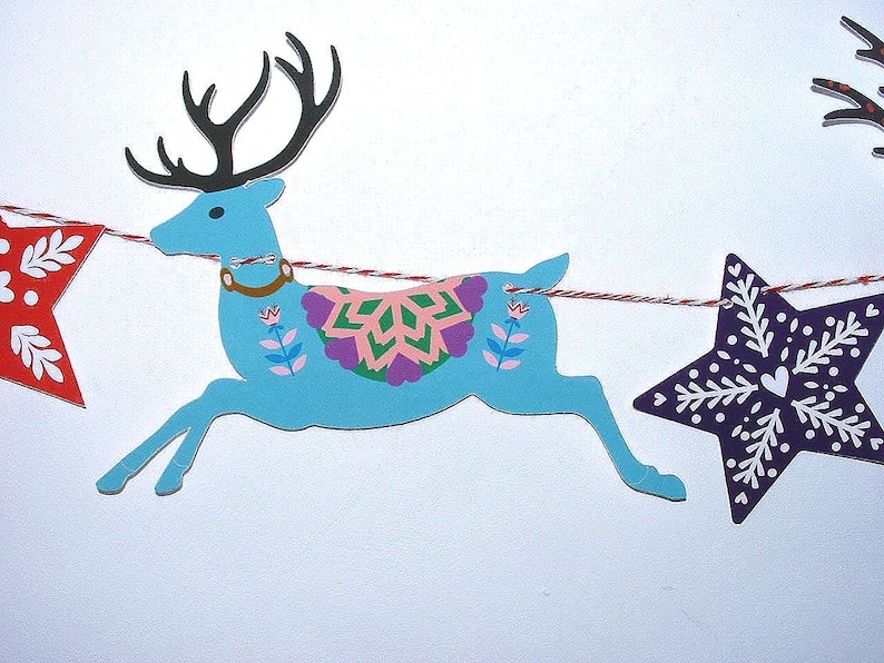 May include: A decorative garland featuring a light blue reindeer cutout with black antlers and a colorful floral saddle, flanked by a red star cutout with white leaf patterns and a dark purple star cutout with white snowflake designs on a string.