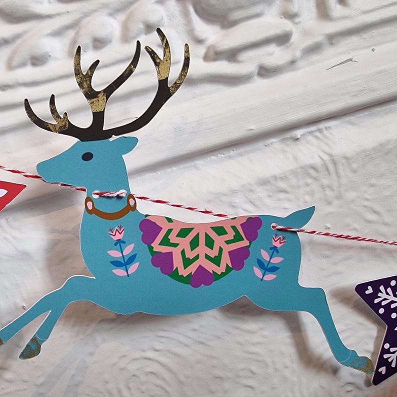 May include: A light blue paper reindeer cutout featuring black antlers with gold accents, a brown collar, and a colorful floral and geometric pattern on its side. The reindeer is strung on a red and white twisted cord as part of a decorative banner.