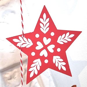 May include: A red star-shaped ornament featuring a white printed pattern of central heart shapes, surrounding dots, and leafy sprigs extending toward each point. The ornament is suspended by a piece of red and white twisted twine string.
