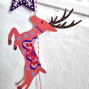 May include: A flat, die-cut paper ornament shaped like a leaping deer in shades of coral pink, featuring dark brown antlers, light blue star patterns, and a central red floral motif, suspended by a red and white twisted string from a purple star.