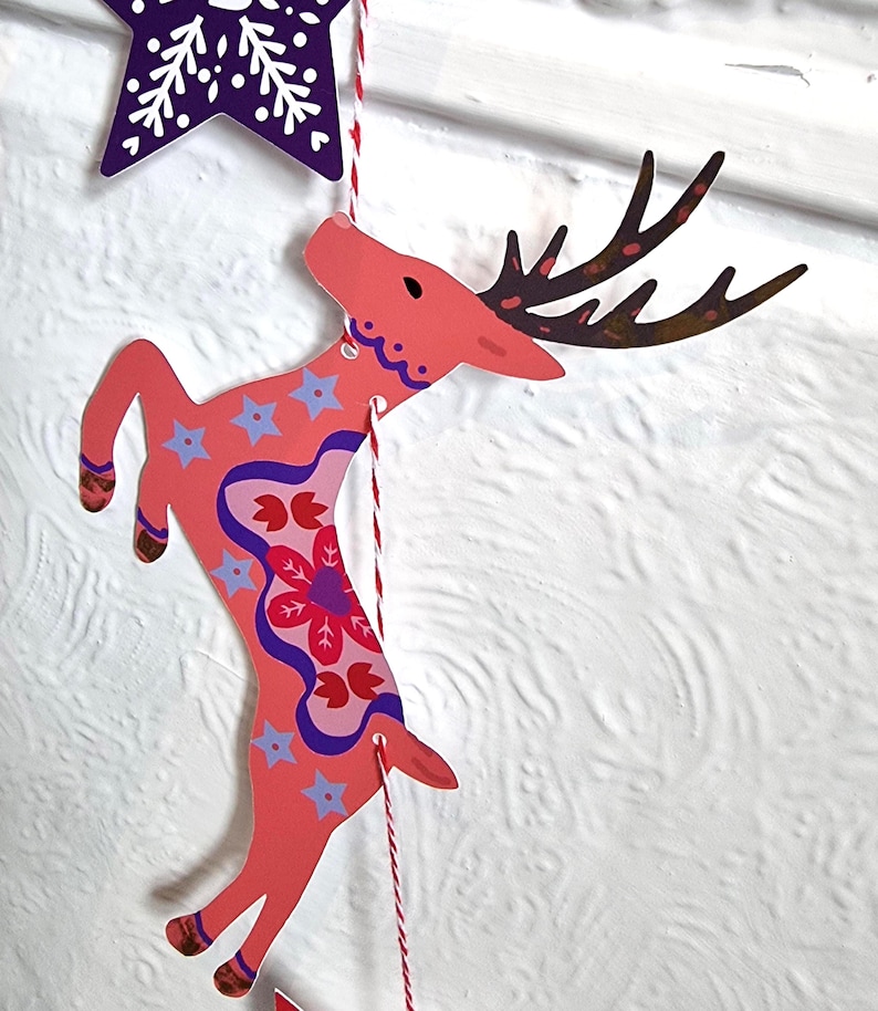 May include: A flat, die-cut paper ornament shaped like a leaping deer in shades of coral pink, featuring dark brown antlers, light blue star patterns, and a central red floral motif, suspended by a red and white twisted string from a purple star.