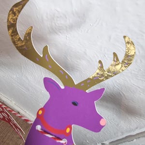 May include: A flat, die-cut paper reindeer ornament featuring a vibrant purple head and body with a pink nose. The reindeer has large, metallic gold antlers with purple accents and a red collar detail. A red and white baker's twine is threaded through.
