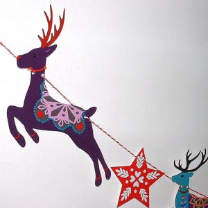 May include: A decorative garland featuring a purple reindeer with red antlers and a pink snowflake pattern, a red star with white floral details, and a blue reindeer with black antlers, all strung together on a red and white twisted twine cord.