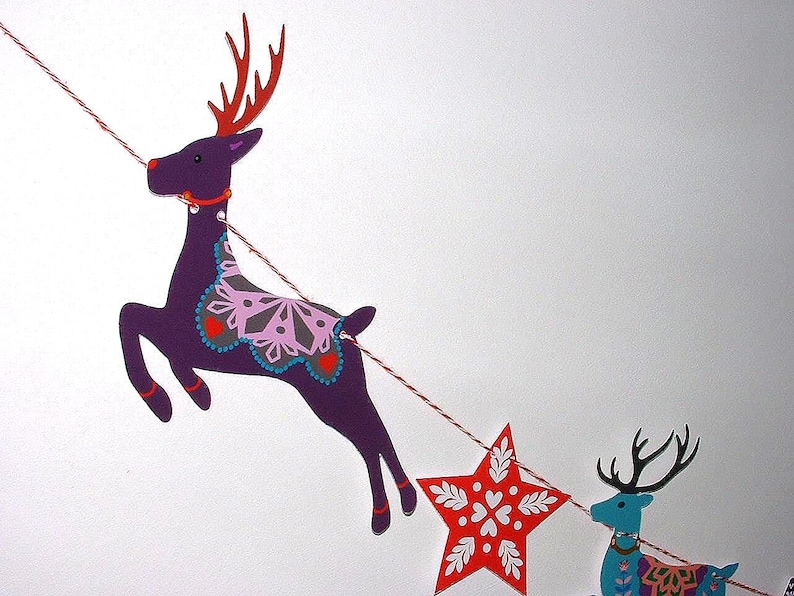 May include: A decorative garland featuring a purple reindeer with red antlers and a pink snowflake pattern, a red star with white floral details, and a blue reindeer with black antlers, all strung together on a red and white twisted twine cord.