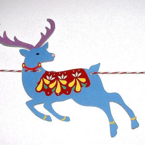 May include: A blue reindeer cutout with purple antlers, a red collar, and a red blanket featuring yellow and white floral patterns. The reindeer is suspended on a red and white twisted string, positioned between a red star and a purple star.