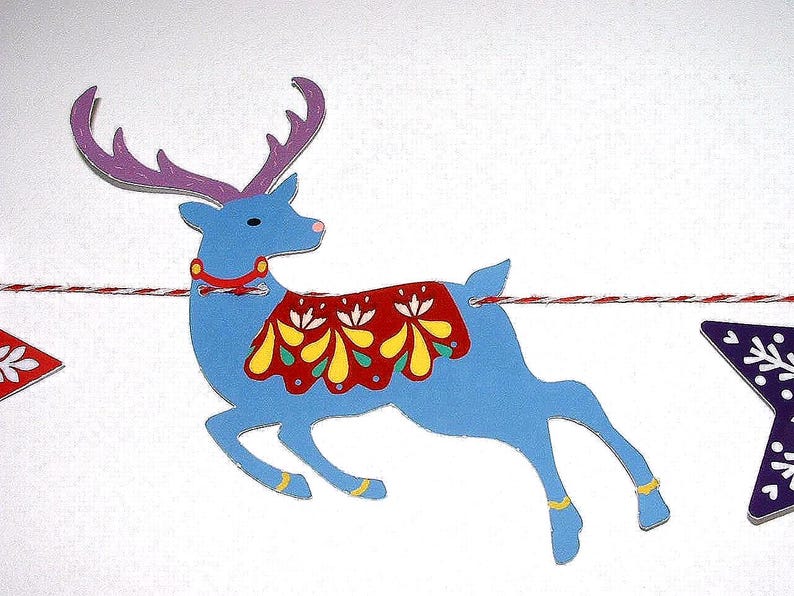 May include: A blue reindeer cutout with purple antlers, a red collar, and a red blanket featuring yellow and white floral patterns. The reindeer is suspended on a red and white twisted string, positioned between a red star and a purple star.