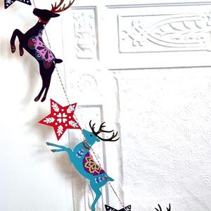 May include: A decorative garland featuring alternating cutouts of leaping reindeer and stars. The reindeer are colored in dark purple and bright blue with intricate floral patterns, while the stars are red with white snowflake-like designs.