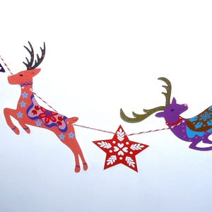 May include: A decorative garland featuring alternating paper cutouts of leaping reindeer and stars strung on red and white twine. The reindeer are colored in shades of coral and purple with intricate floral and snowflake patterns in blue and white.