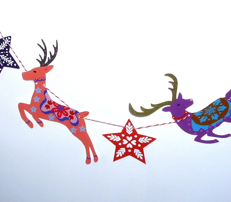 May include: A decorative garland featuring alternating paper cutouts of leaping reindeer and stars strung on red and white twine. The reindeer are colored in shades of coral and purple with intricate floral and snowflake patterns in blue and white.