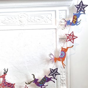 May include: A decorative garland featuring alternating paper cutouts of leaping reindeer and stars. The reindeer are colored in shades of blue, orange, and pink with intricate floral patterns. The stars are dark blue and red with white snowflake designs.