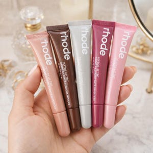 May include: Five tubes of Rhode Peptide Lip Tint in various shades of pink, brown, and white are held in a hand. Each tube has a matching colored cap and the brand name "rhode" printed on the front. The tubes are labeled with product information.
