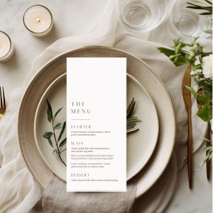 May include: A vertical white menu card with black serif text titled THE MENU, listing sections for STARTER, MAIN, and DESSERT. The menu features items like creamy burrata, grilled filet, and layered cake, centered on a speckled ceramic plate.
