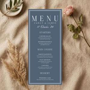 May include: A vertical rectangular menu card in dusty blue with white text reading MENU, CARLY & JAMES, 17 October 26, and a list of food items including Caesar salad, sourdough rolls, baked haddock, filet mignon, lasagna, and wedding cake.