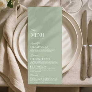 May include: A sage green rectangular menu card featuring white text that reads The MENU AUGUST 22 2026, listing Caesar Salad, Chicken Piccata, Filet Mignon, and Vanilla & Berry Cake, centered on a stack of white ceramic plates with a beige napkin.