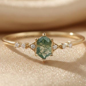 Op de afbeelding: Een gouden ring met een zeshoekige groene mosagaat edelsteen in het midden. De ring is versierd met kleine, ronde, heldere edelstenen aan weerszijden van de centrale steen. De band is dun en goudkleurig, met de tekst "AureviaGems" zichtbaar.