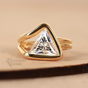 May include: A gold ring with a triangular, clear gemstone. The ring's band is a simple, smooth gold, and the gemstone is set in a gold frame. The ring is on a neutral-toned surface. The Diamond Storie logo is in the upper right corner.