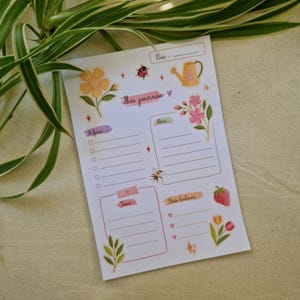 May include: A white paper daily planner sheet featuring the text Ma journée, A faire, Notes, and Petits bonheurs. The design includes illustrations of a yellow flower, a ladybug, a yellow watering can, pink flowers, a bee, a strawberry, and tulips.