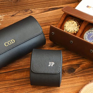 Peut inclure: Un étui à montres et un coffret à montres en cuir noir avec des initiales dorées. Un coffret à montres en cuir marron contient trois montres avec des cadrans dorés, argentés et bleus. Sur une surface en bois.
