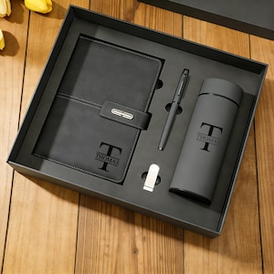 Puede incluir: Un juego de regalo gris que contiene un cuaderno, un bolígrafo, una botella de agua y un clip. El cuaderno tiene un cierre de solapa y el nombre "THOMAS" impreso. La botella de agua también tiene el nombre "THOMAS" impreso. Los artículos están en una caja negra.