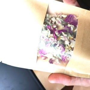 Op de afbeelding: Een rechthoekig zakje van bruin papier met een doorzichtig plastic venster aan de voorzijde. In het venster is een mengsel van gedroogde botanische elementen zichtbaar, waaronder levendige paarse en witte bloemblaadjes en groene blaadjes.
