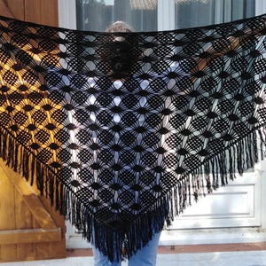 Peut inclure: Un châle noir en crochet fait à la main, avec un motif géométrique et une bordure à franges. Le châle est tenu, mettant en valeur sa forme triangulaire et son design ajouré. Les franges pendent le long du bord inférieur.