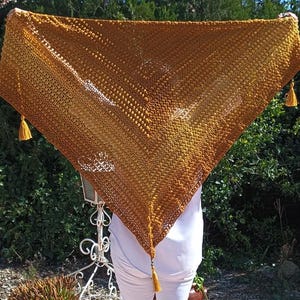 Peut inclure: Un châle triangulaire fait à la main dans des tons or et marron, avec un tissage ouvert texturé. Le châle a un gland à chaque pointe. L'article est présenté sur un fond de feuillage vert.